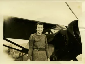 A woman wearing a belted dress stands smiling in front of an old-fashioned propeller airplane. Parts of the airplane’s wing and engine are clearly visible beside her. The photo is sepia-toned, giving it a vintage appearance.