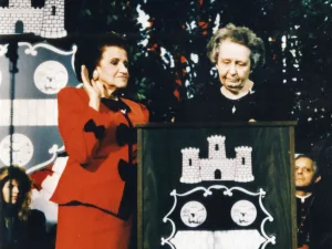 Two women stand at a podium decorated with a black-and-white castle emblem. One woman, dressed in a red suit, raises her right hand, while the other woman looks down at the podium. Several seated people are visible in the background.
