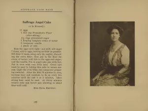 Two open book pages; the left page displays a recipe titled Suffrage Angel Cake with a list of ingredients and step-by-step instructions in black text on white paper. The right page features a sepia-toned photograph of a woman from the early 1900s, seated and facing forward, dressed in period attire with a high-collared blouse and long skirt.
