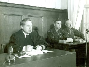 A judge in a dark robe sits at a wooden desk with a gavel and several papers, facing forward. Two military officers in formal uniforms are seated beside him at the same bench. The background features wood paneling and a window with curtains.