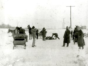 Black-and-white photograph of a group of people gathered on a snow-covered rural road. Horses and sleds are visible in the background. Several individuals are standing or kneeling around a person lying on the ground, under an overcast sky, indicating a possible emergency or incident.