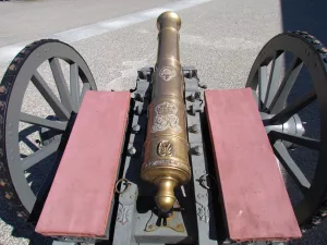 A close-up view of an old brass cannon with ornate engravings, mounted on a gray wooden carriage with two large spoked wheels and red padded seats on each side. The cannon is outdoors on a paved surface.