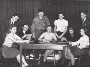 A black-and-white photo of eight women and two men gathered around a wooden table, some seated and some standing, looking at papers with attentive expressions. The background features curtains.