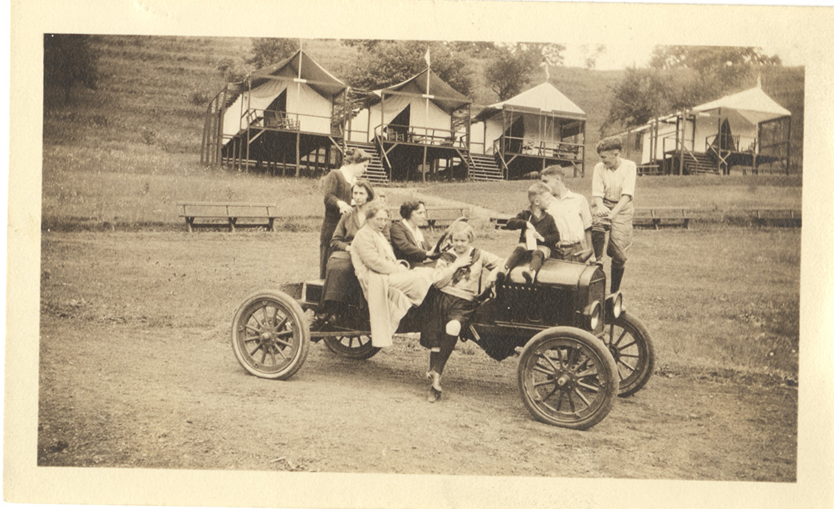 Kids and adults pile on a vintage car in front of a row of tents on platforms, c. 1920. General Photograph Collection, Detre Library & Archives at the History Center.