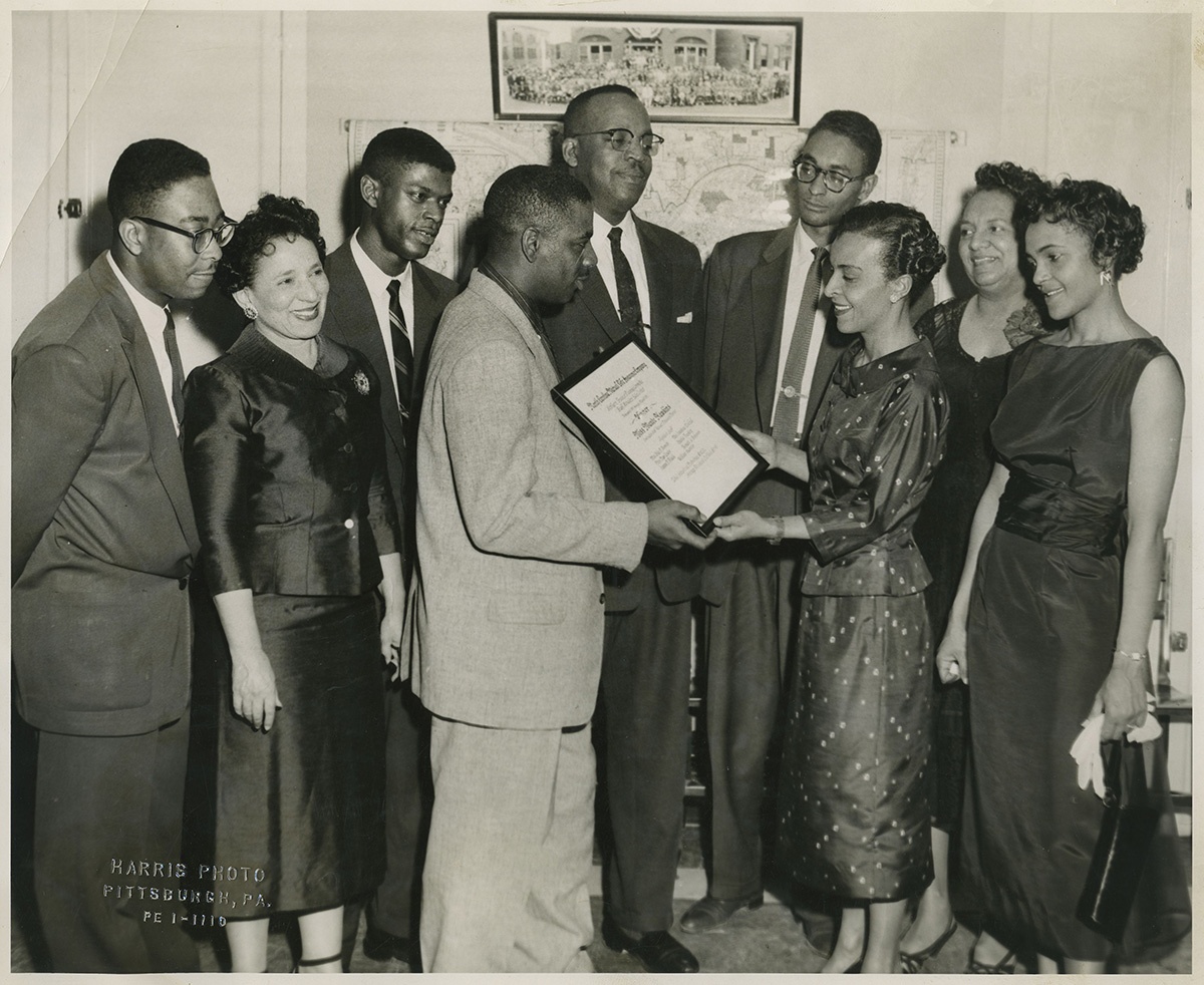 Group portrait of four women, and five men, including one wearing light colored cross hatch patterned suit presenting certificate inscribed "North Carolina Mutual Life Insurance Company...winner Miss Maude Hawkins..." to woman wearing dark suit with small flower or dot pattern, in interior with map and large group photograph on wall, another version, c. 1957. Maude Y. Hawkins Photographs, MSQ 171, Detre Library & Archives at the History Center. © Carnegie Museum of Art, Charles “Teenie” Harris Archive.