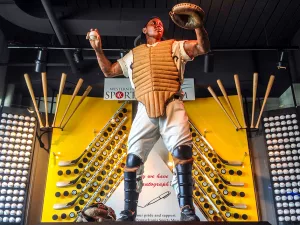 A life-sized statue of a baseball catcher in full protective gear stands in front of a display with crossed bats and baseballs, posed as if ready to throw. The background shows a yellow and white wall with sports-themed decorations.
