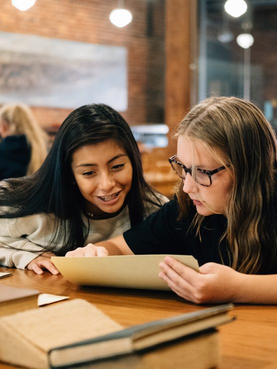 Two girls sitting at a wooden table in a library, smiling and looking at a tablet together. Books are scattered on the table, and several people are visible in the background among bookshelves.