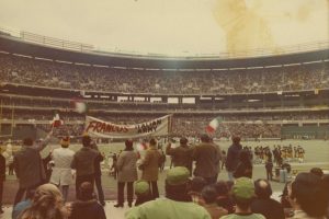 A crowded football stadium with fans in the stands, many holding Italian flags and a large banner that reads “Franco’s Italian Army”; the atmosphere is lively as spectators cheer and watch the game.