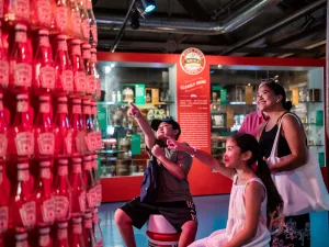 A boy, a girl, and a woman smiling and pointing excitedly at a wall display of stacked ketchup bottles inside a colorful museum exhibit; the background includes historical items and a red information board.