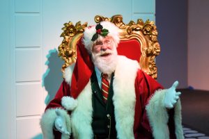 A person dressed as Santa Claus, wearing a red suit with white fur trim and a holly brooch, sits on an ornate golden throne. Santa is smiling and gesturing with both hands.