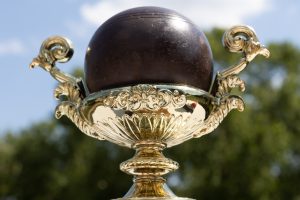 A gold trophy with ornate handles and a dark bocce ball on top is displayed outdoors. The plaque reads: Italian American Program Bocce Tournament Champions, Senator John Heinz History Center. Trees and a partly cloudy sky are visible in the background.