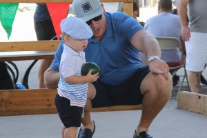A man crouches next to a small child holding a bocce ball, guiding him on how to play on an outdoor bocce court. Several other people and colorful flags are visible in the background.