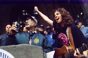 A man speaks into a handheld radio at a podium while a woman standing next to him raises her fist and smiles, holding a guitar. They are surrounded by people at a lively rally.