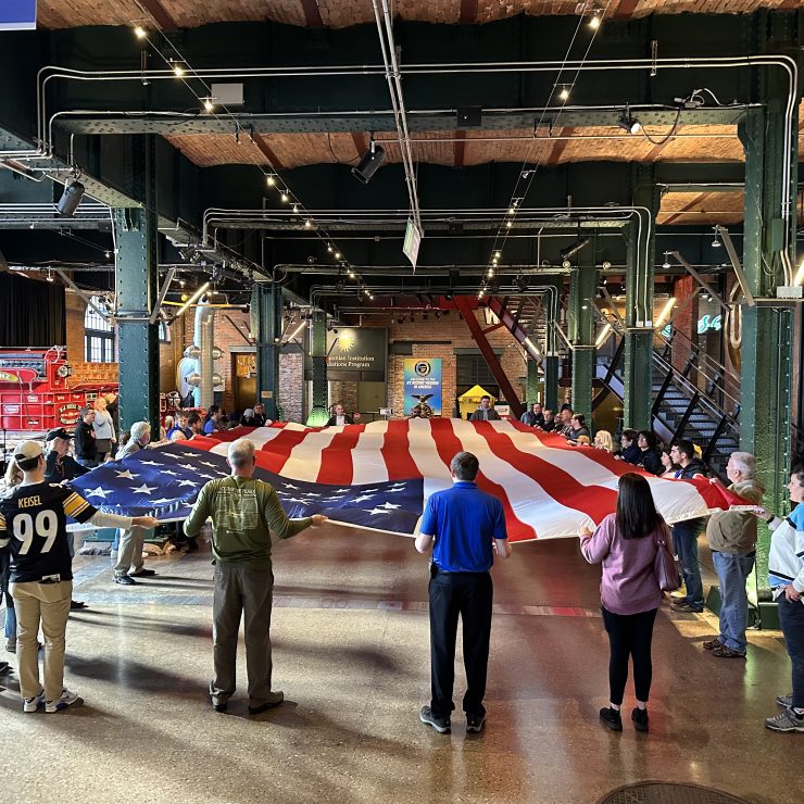 A group of people stand in a circle inside an industrial-style building, each person holding the edge of a large American flag that is stretched out between them during a Flag Day folding event. Various informational signs and museum displays are visible in the background.