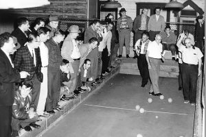 Italian men play bocce while a crowd of other men watch.