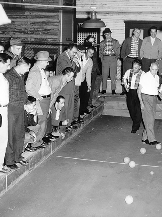 Italian men play bocce while a crowd of other men watch.