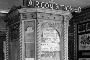 Black-and-white photo of vintage theater ticket booth with ornate woodwork, AIR CONDITIONED sign, admission info, film posters. *AI generated alt text