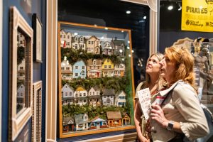 Two women stand side by side in a museum exhibit, both looking at colorful house-themed artwork. One woman holds a brochure and a phone. Behind them are additional framed artworks, museum artifacts, and a yellow Pittsburgh Pirates banner displayed on the wall.