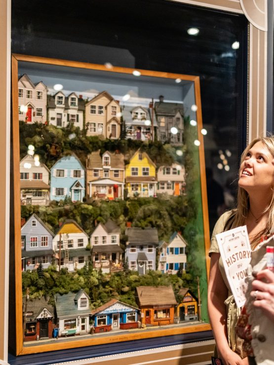 Two women stand side by side in a museum exhibit, both looking at colorful house-themed artwork. One woman holds a brochure and a phone. Behind them are additional framed artworks, museum artifacts, and a yellow Pittsburgh Pirates banner displayed on the wall.