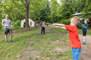 A young boy in a red shirt swings a stick with a hoop on it, while two other children and an adult watch during a homeschool activity in a grassy, wooded yard near a small yellow house.
