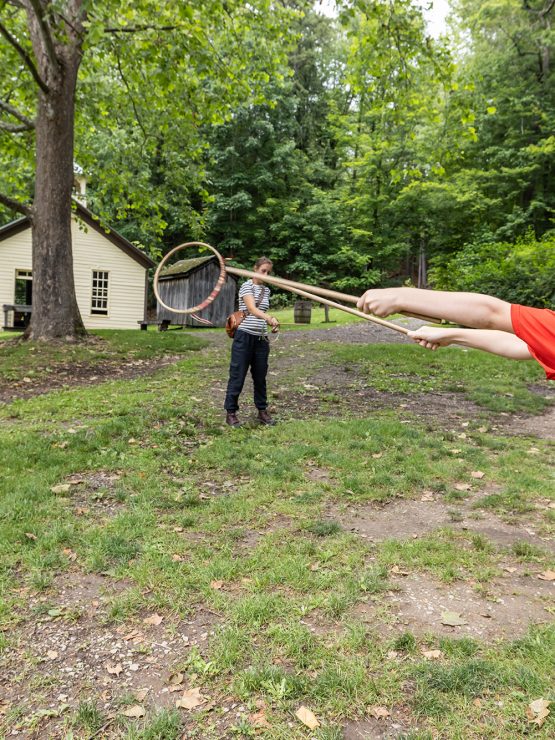 A young boy in a red shirt swings a stick with a hoop on it, while two other children and an adult watch during a homeschool activity in a grassy, wooded yard near a small yellow house.