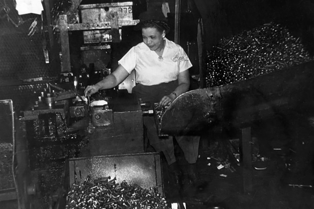 A woman stands at a factory workstation operating machinery, surrounded by large bins filled with metal bullet casings or ammunition. She is focused on her task, wearing work clothes and using both hands to manipulate the equipment. The setting features industrial lighting and additional factory equipment in the background, evoking a wartime assembly line environment.