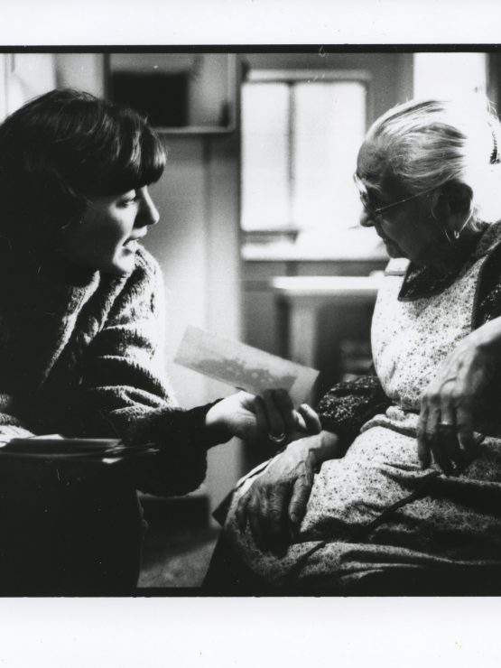 A young person sits next to an elderly woman, holding up a piece of paper for her to see. They are engaged in conversation in a cozy room with soft sunlight streaming through the window. The photo is black and white.