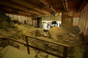 Excavation site covered by a wooden roof with exposed support beams; ancient stone ruins and scattered artifacts partially embedded in the soil; several visitors standing behind a glass barrier observe the archaeological dig area.