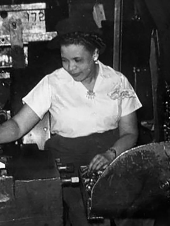 A woman wearing a white blouse operates machinery in a factory setting, standing next to metal equipment and a large container filled with metal rods or parts; the image depicts an industrial and historical environment.