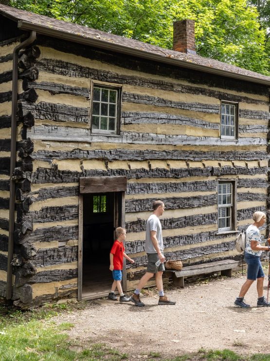 A family with children walks toward the entrance of an old log cabin, surrounded by green trees on a sunny day.