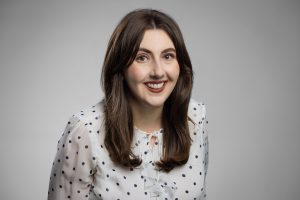 A woman with long brown hair, wearing a white blouse with black polka dots, smiles at the camera against a plain grey background.