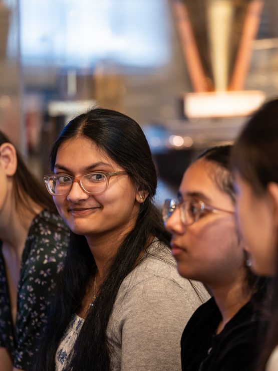 Four young women with long hair and glasses sit side by side indoors during a museum group tour. One in the center smiles at camera. *AI generated alt text