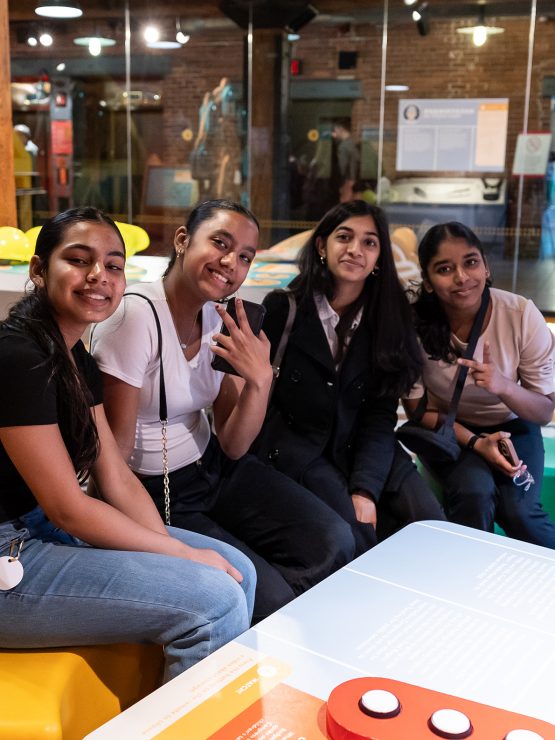 Four young women sit together smiling in a modern US history museum with interactive exhibits and bright seating. *AI generated alt text