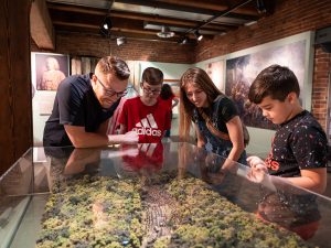 A man and three children observe a glass-covered forest landscape diorama in a history museum, with panels and images behind them. *AI generated alt text
