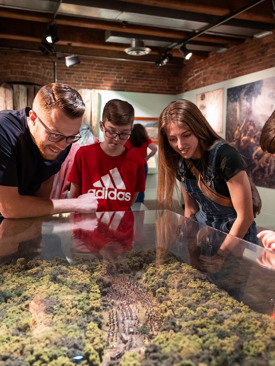 A man and three children observe a glass-covered forest landscape diorama in a history museum, with panels and images behind them. *AI generated alt text