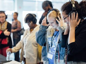 A group of visitors at a history museum wear headphones and listen closely, some interacting with a display table in a softly lit room. *AI generated alt text