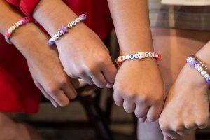 Three children’s wrists with beaded bracelets spelling “KIDS,” “ARE,” and “SAFE.” They wear red and tan clothing. *AI generated alt text