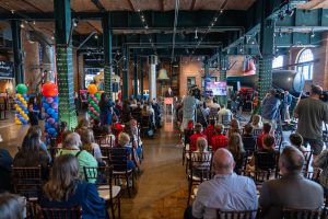Visitors seated in rows face a stage in a brick-walled hall with exposed beams; balloons and AV equipment present during a podium talk. *AI generated alt text