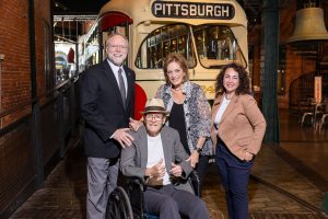 Four adults, including one in a wheelchair, pose in front of a vintage Pittsburgh streetcar at the Kamin Museum with exhibits behind. *AI generated alt text