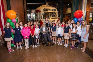 A group of schoolchildren and two adults pose in front of a vintage Pittsburgh trolley in a museum, with colorful balloon decorations. *AI generated alt text
