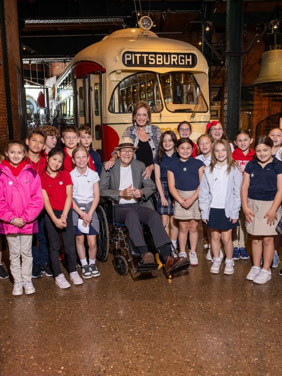 A group of schoolchildren and two adults pose in front of a vintage Pittsburgh trolley in a museum, with colorful balloon decorations. *AI generated alt text