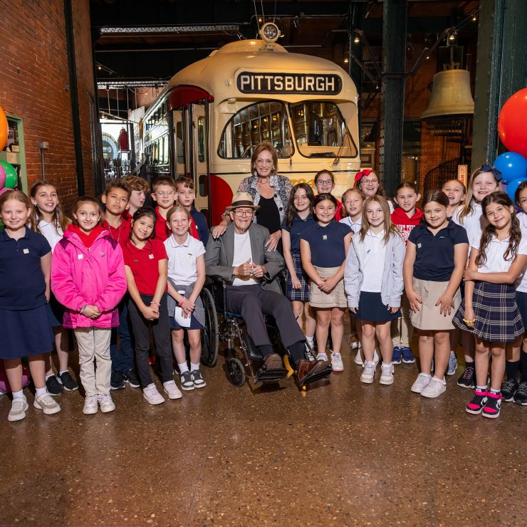 A group of schoolchildren and two adults pose in front of a vintage Pittsburgh trolley in a museum, with colorful balloon decorations. *AI generated alt text