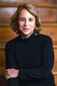 A middle-aged woman with short, wavy blonde hair wearing a black long-sleeve top sits with her arms gently crossed and smiles softly while seated in front of blurred wooden steps at the Smithsonian. AI generated alt text