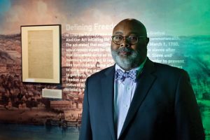 A man with glasses and a gray beard in a suit and bow tie stands before an exhibit titled Defining Freedom with a framed document. *AI generated alt text