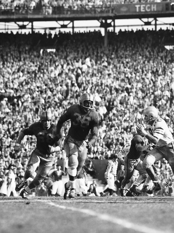 Black-and-white photo of a crowded football stadium with players in motion; one player prepares to throw the ball during a game. *AI generated alt text