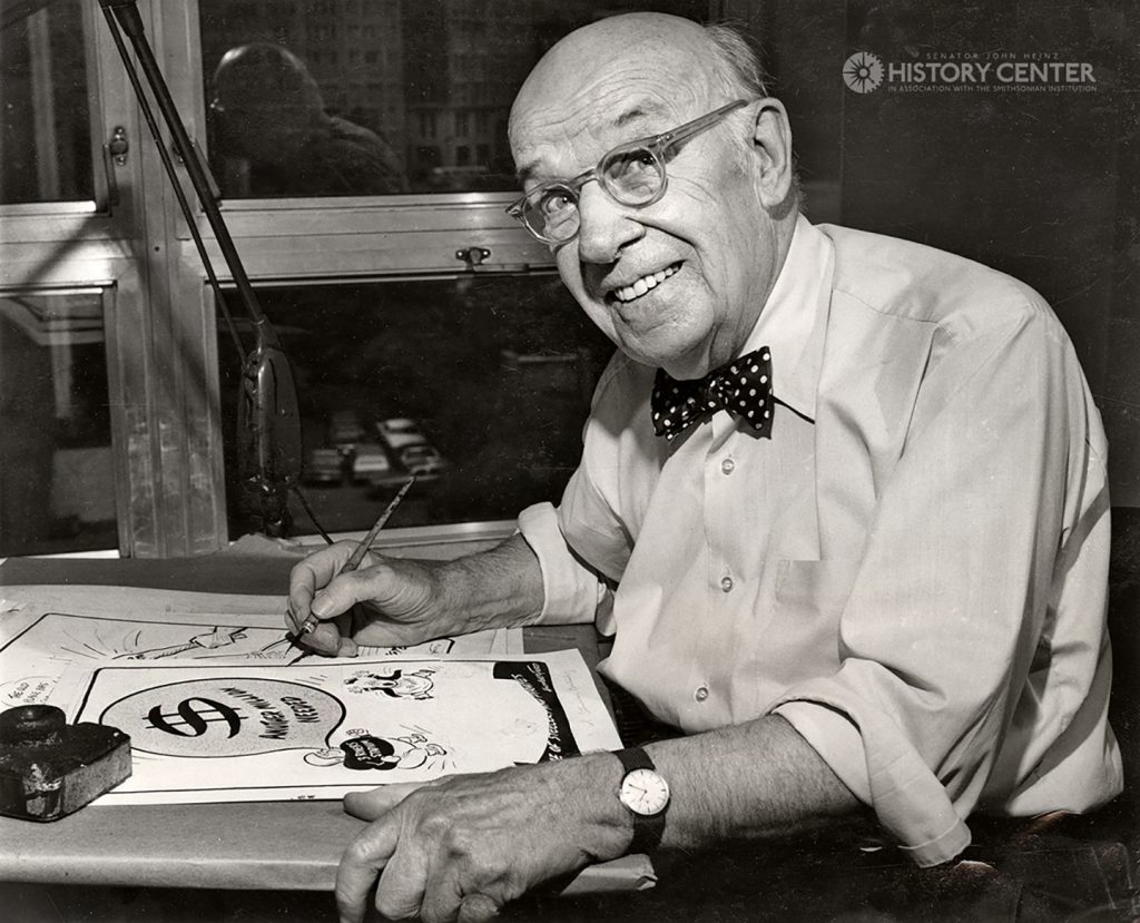 An older man with glasses, bow tie, and watch draws a cartoon with sketches and a large dollar sign at a desk. History Center watermark. *AI generated alt text
