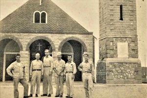 Six men in military uniforms stand in front of a stone church with arched doorways and a cross above the entrance, near a tall stone tower with a gated door. The photograph is sepia-toned and appears aged.