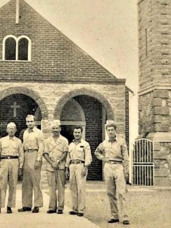 Six men in military uniforms stand in front of a stone church with arched doorways and a cross above the entrance, near a tall stone tower with a gated door. The photograph is sepia-toned and appears aged.