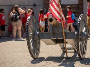 A historic cannon with an American flag stands outdoors. Nearby, children in red shirts and masks visit during a Fort Pitt field trip. *AI generated alt text