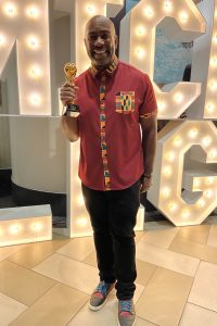A man in a red shirt with colorful trim holds a World Cup-like trophy indoors on tiled flooring, large lit letters behind him. *AI generated alt text